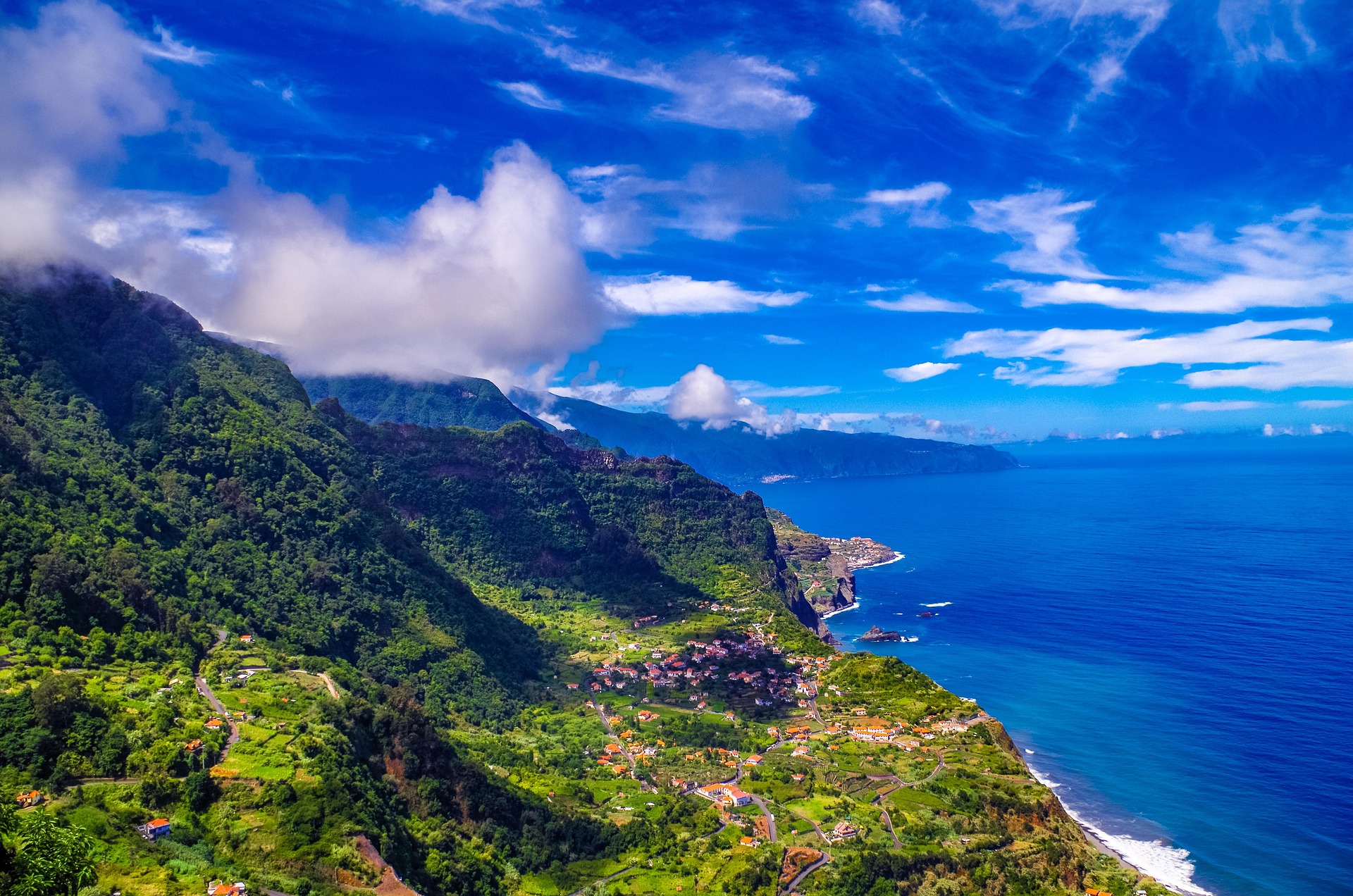 The coast of Madeira in emerald green, against a backdrop of deep blue mostly clear sky and dark blue ocean.